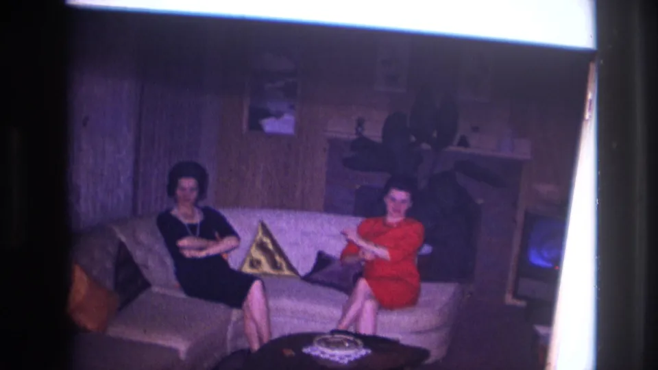 1967 Ottawa home-movie frame showing two women seated on a sofa in a private living room, one in black with pearls and one in red.