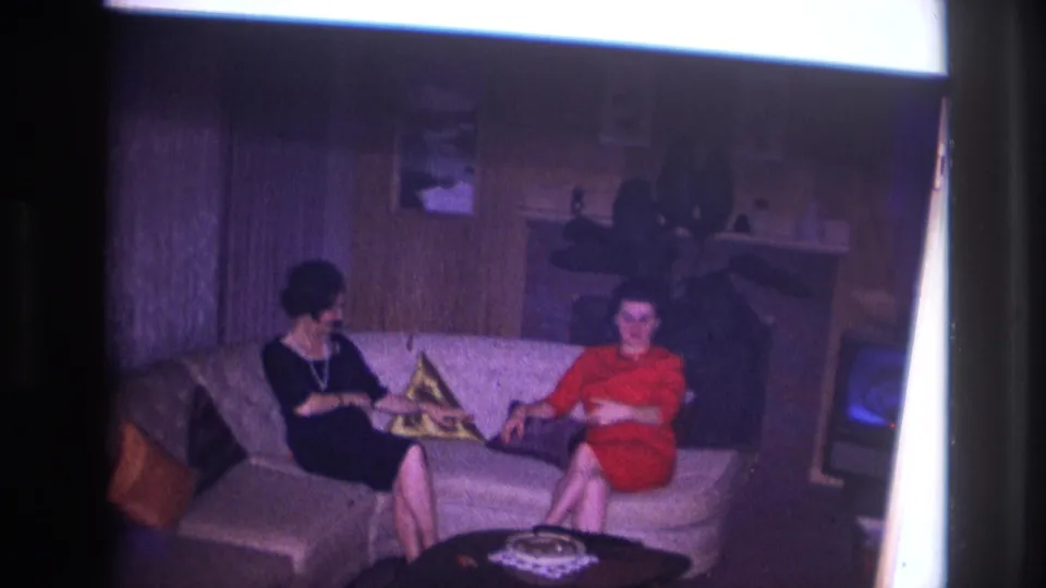 1967 Ottawa home-movie frame of two women on a living-room sofa, one in a black dress turning toward a woman in red.
