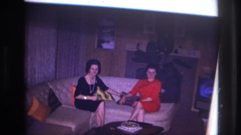 1967 Ottawa home-movie frame showing two women seated in a private living room with a coffee table, pillows, and wood paneling.