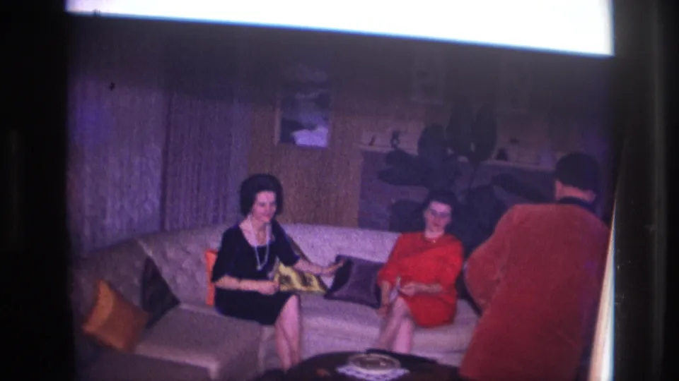 1967 Ottawa home-movie frame with two women on a sofa and a man in a red sweater entering at the right edge of the private room.