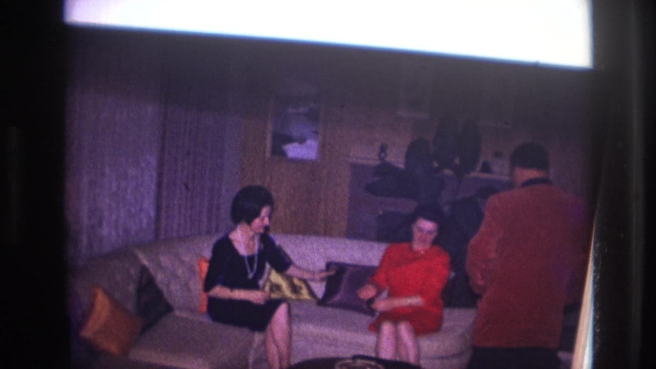 In a 1967 Ottawa home-movie frame, two women sit on a sofa as a man in a red jacket stands beside them in a private living room.