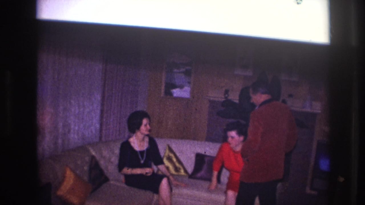 A woman in black and a woman in red sit on a sofa as a man stands in front of them.