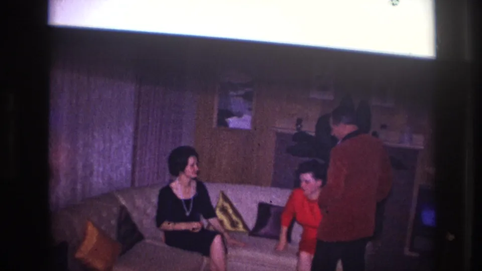 The Ottawa living room scene shows two women on a sofa, one in black and one in red, with a man standing close by during a social visit.