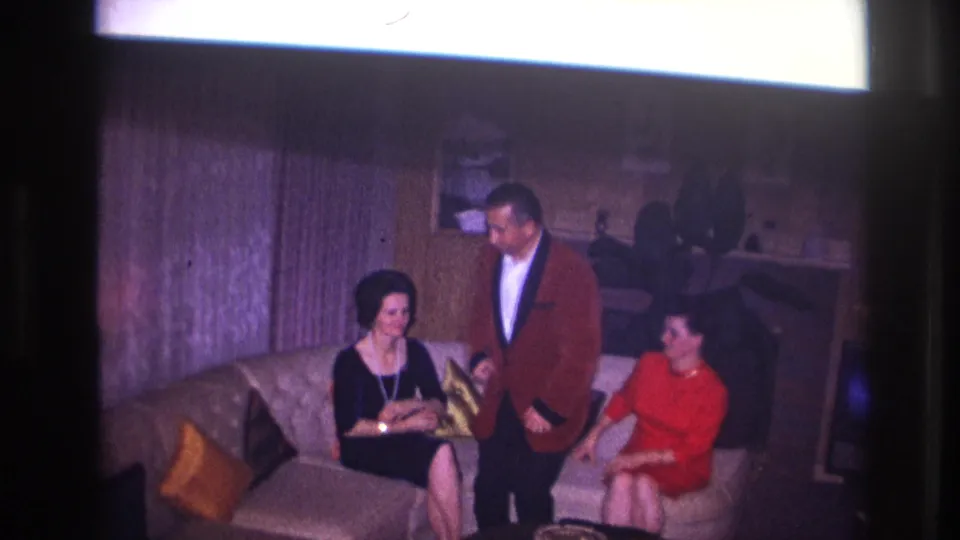In this 1967 home-movie frame, the man stands at the sofa between two women in a private Ottawa living room with mid-century decor.