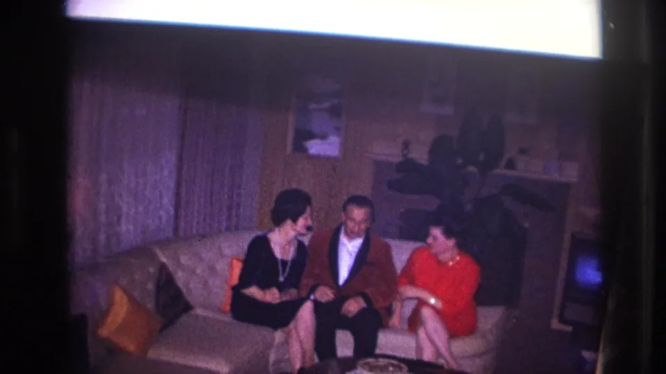 Three adults sit together on a sofa in the 1967 Ottawa home-movie scene, framed by wood paneling, cushions, and a potted plant.