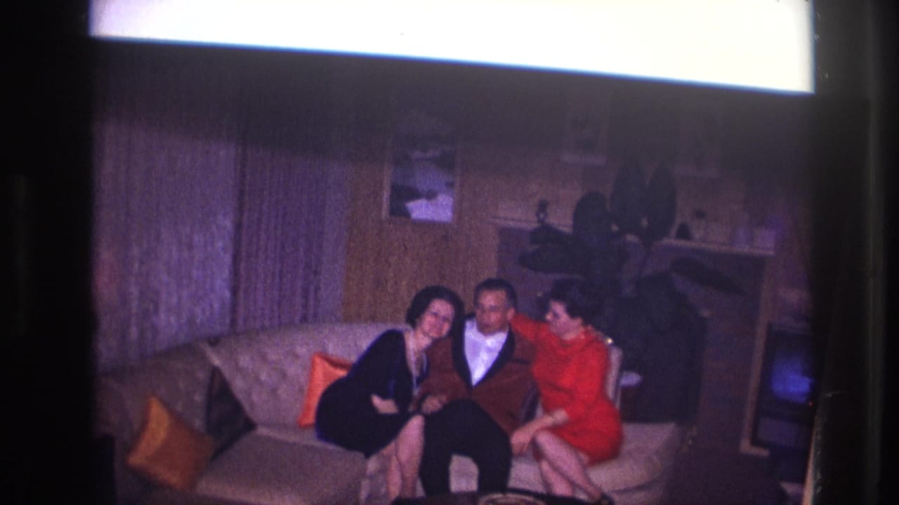 Two women lean toward a seated man on a sofa in a living room.