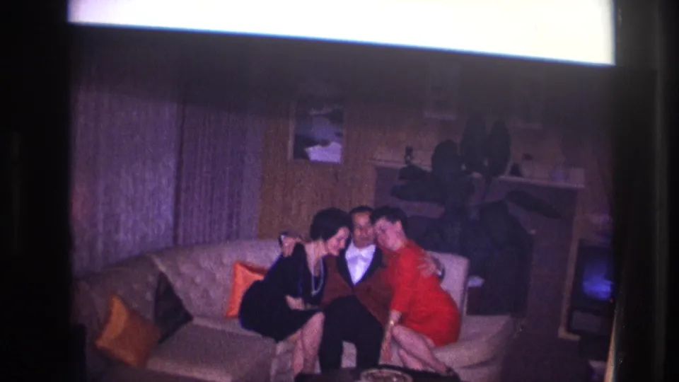 The adults draw closer on the sofa, framed by wood paneling, curtains, wall art, and a faint television glow.