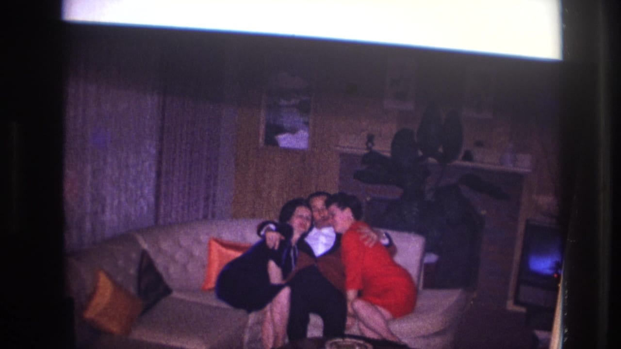 Ottawa 1967 home-movie frame showing two women leaning in toward a man in a tuxedo on a light couch in a wood-paneled living room.