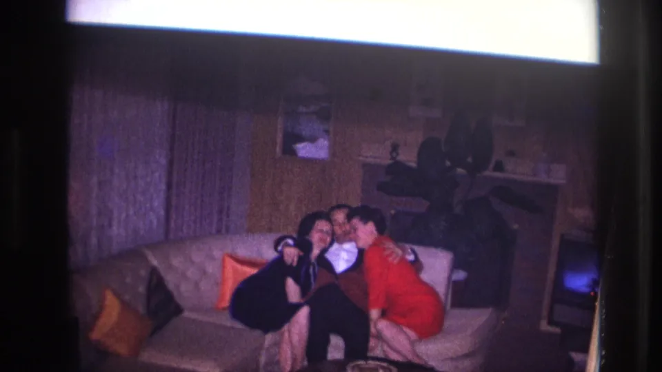 Ottawa 1967 home-movie frame showing two women leaning in toward a man in a tuxedo on a light couch in a wood-paneled living room.