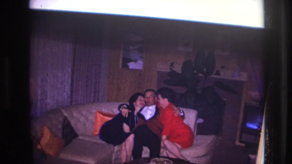 Ottawa 1967 frame with a man in formalwear seated between two women on a couch in a dim wood-paneled living room.