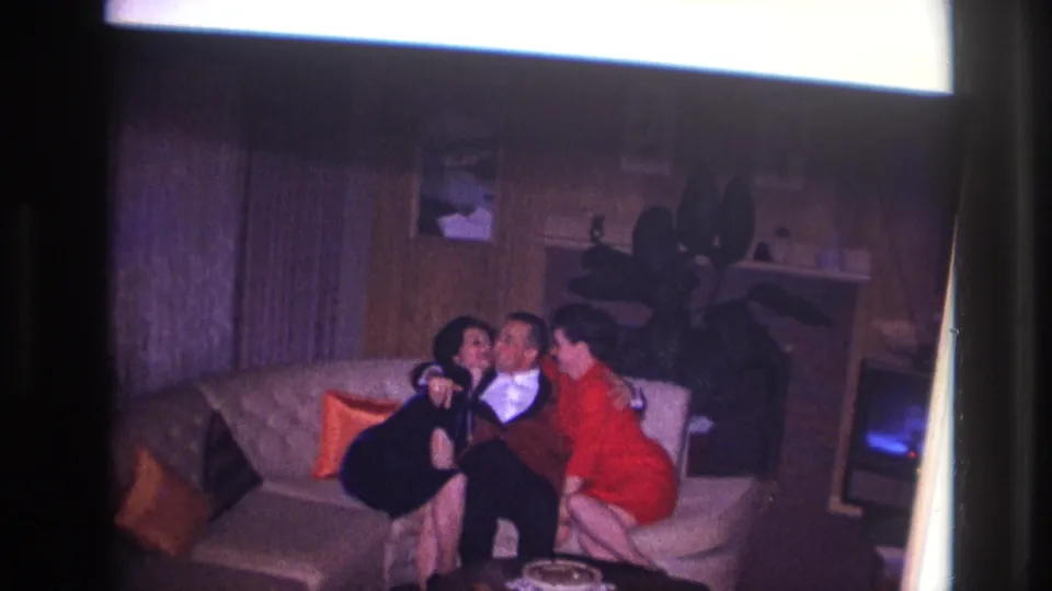 1967 Ottawa home-movie frame of a seated man between two women on a couch, with a fireplace area and small television nearby.