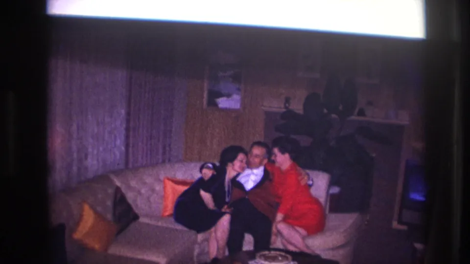 Ottawa 1967 8mm frame showing two women close to a man in a tuxedo on a couch, with wood paneling and party-like posing.