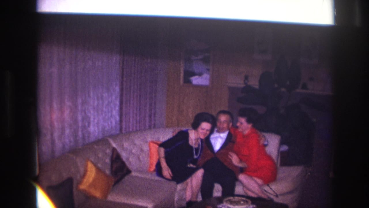 In 1967 Ottawa home-movie footage, a man sits between two women on a light couch beside purple curtains and pillows.