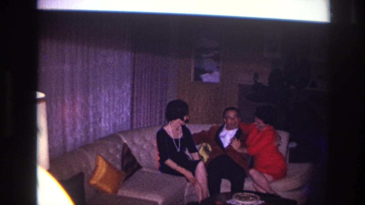 Two women and a man sit on a couch while the woman in red leans in.