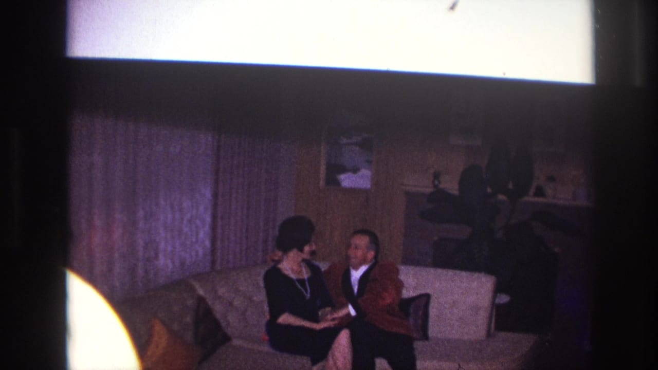 Man and woman seated on a couch in a wood-paneled living room.