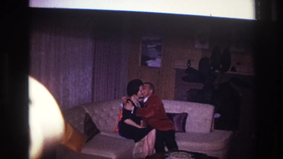 The couple remains close together on the couch, with the man leaning in as the woman faces him in the 1960s interior.