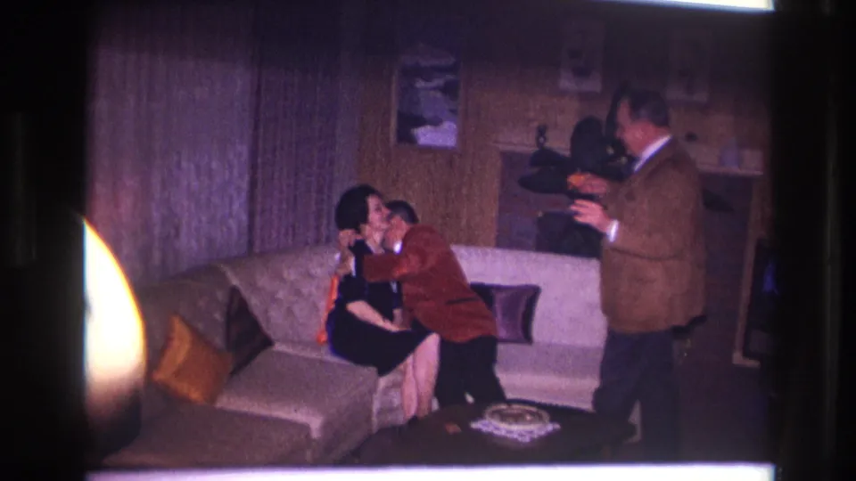 A third man in a suit stands at the right holding a camera as the couple kisses on the couch in the Ottawa home interior.