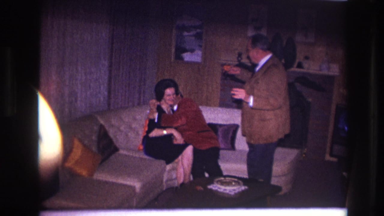 1967 Ottawa home-movie frame of adults celebrating in a wood-paneled living room as two embrace on a sofa and another stands nearby.