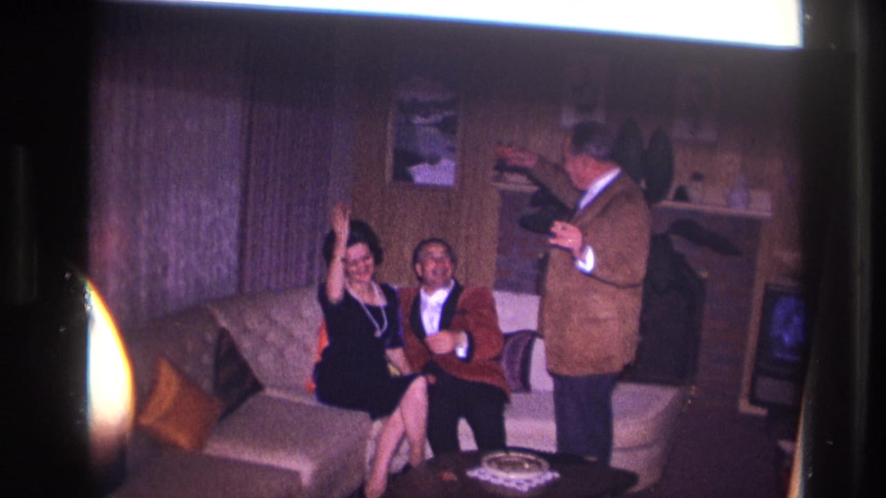 1967 Ottawa home-movie frame with a woman waving from a sofa beside a man in a red jacket as another man stands with a drink.