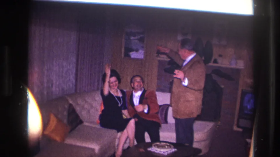 1967 Ottawa home-movie frame with a woman waving from a sofa beside a man in a red jacket as another man stands with a drink.