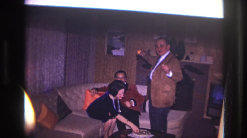 1967 Ottawa home-movie frame showing a woman leaning down near a low table as two men continue the celebration in a paneled room.