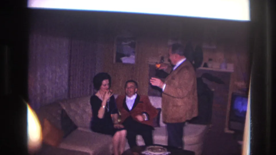 1967 Ottawa home-movie frame of three adults socializing with drinks in a wood-paneled living room with a television nearby.
