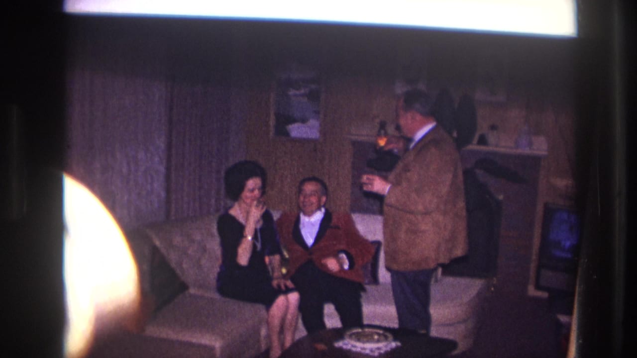 Three adults in a living room as a standing man prepares to light a cigarette.