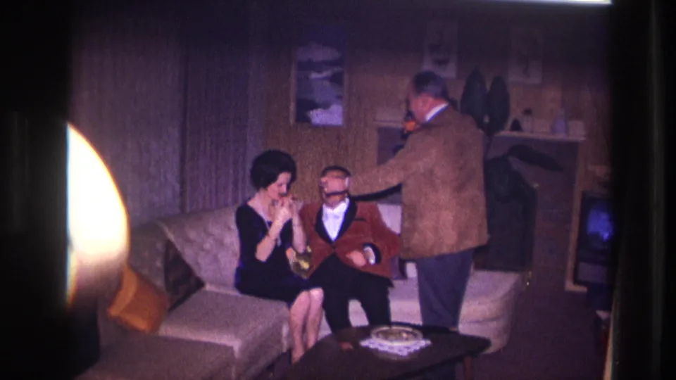 Ottawa 1967 interior frame with a standing man lighting a seated man's cigarette as a woman watches from the sofa.
