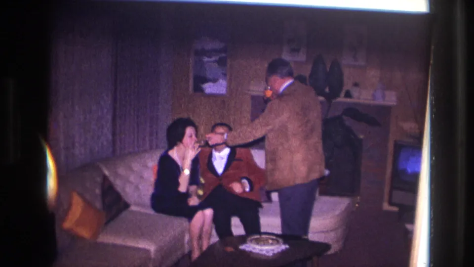 Ottawa 1967 home-movie frame showing a woman on a sofa receiving a light while a seated man looks on.
