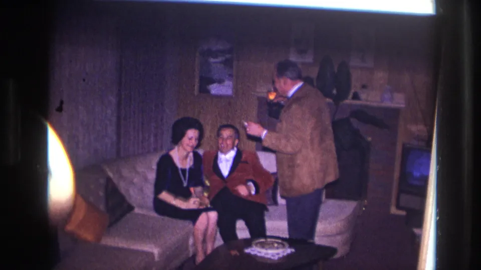 Ottawa 1967 home-movie frame of three adults in a wood-paneled living room, with two seated on a sofa and one standing nearby.