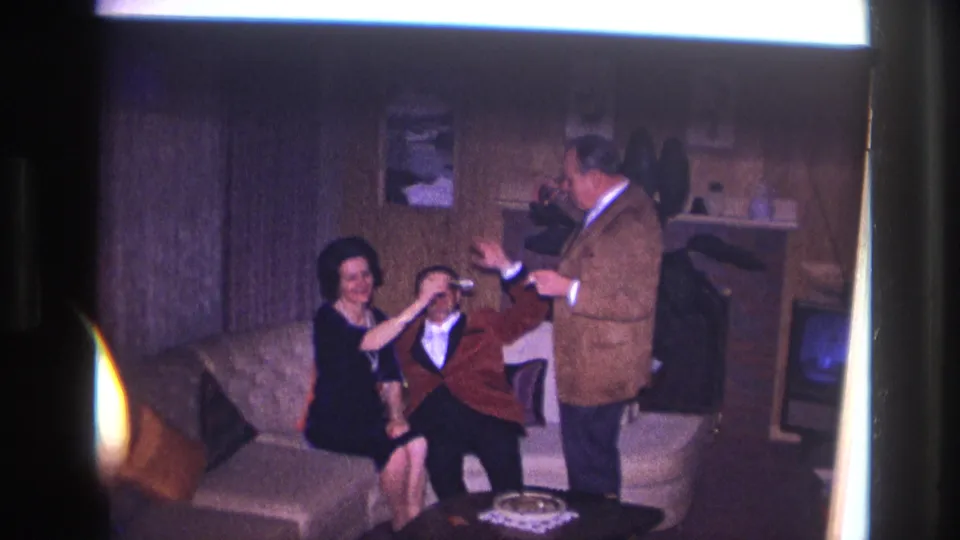 1967 Ottawa home-movie frame of adults joking with drinks beside a sofa and fireplace in a private interior.