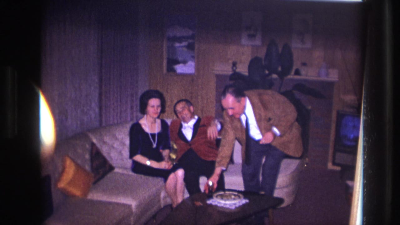A man in a brown jacket pours or places a drink while two adults sit on a sofa.