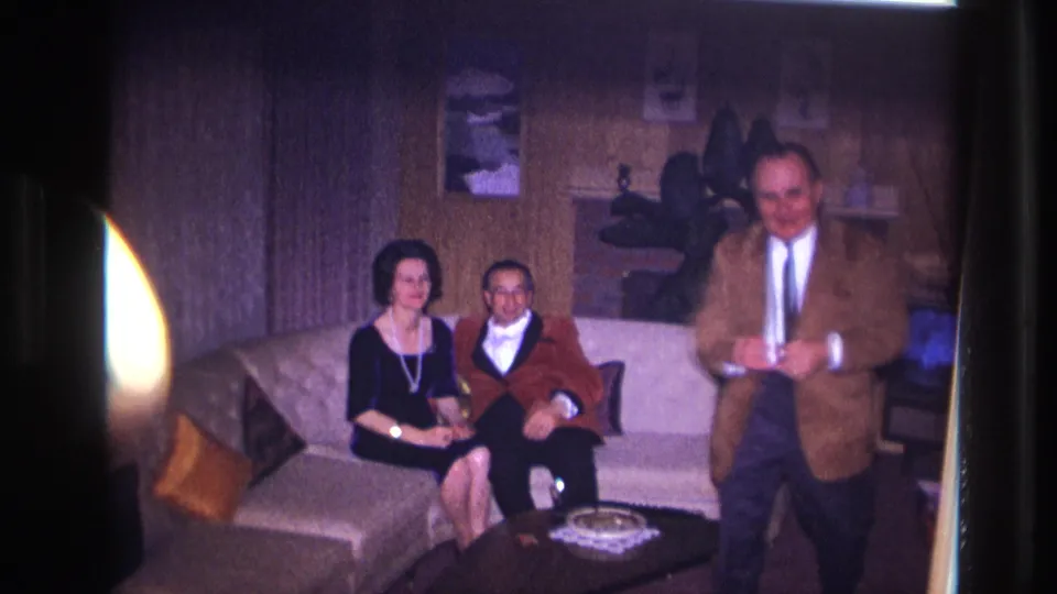The 8mm frame shows two adults seated on a beige sofa in an Ottawa home as a man in a brown jacket passes in front of the camera.