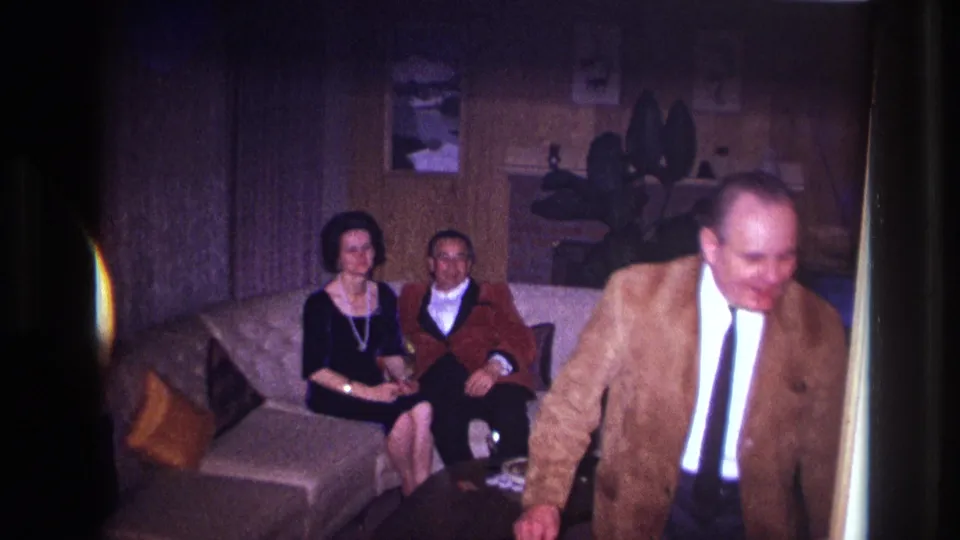 A 1967 Ottawa home-movie frame shows two adults seated in a paneled living room while a smiling man in a brown jacket moves close to the camera.
