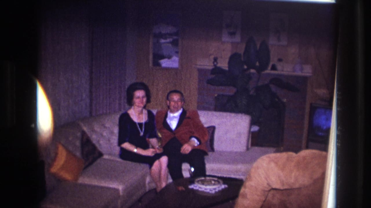 In a 1967 Ottawa home-movie frame, a dressed couple sit on a light sofa with a coffee table, plant, and television nearby.