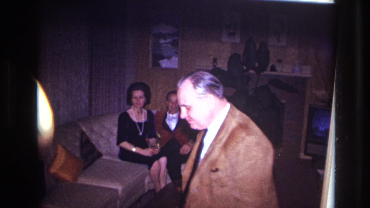 A man in a brown jacket crosses the foreground while the 1967 Ottawa living room couple remains seated on the sofa behind him.