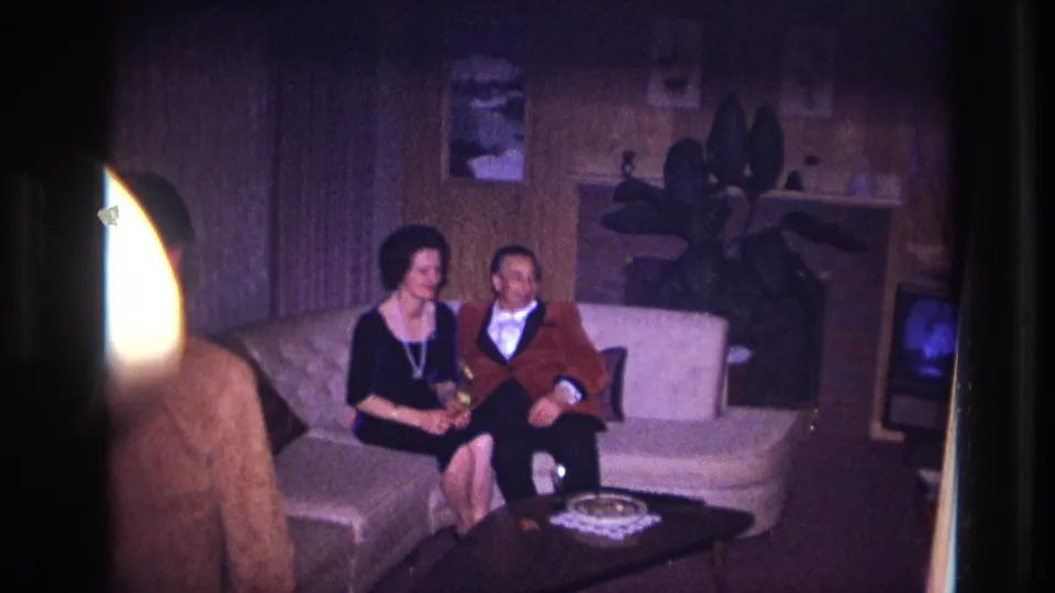 A seated couple in formal clothes look toward the camera as another adult partly enters the foreground of the 1967 Ottawa living room frame.