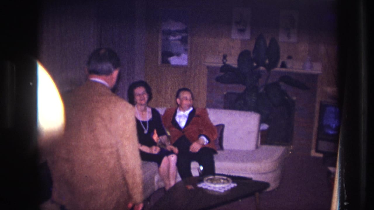 1967 Ottawa home-movie frame showing a man in a brown jacket nearing a formally dressed couple on a white couch.