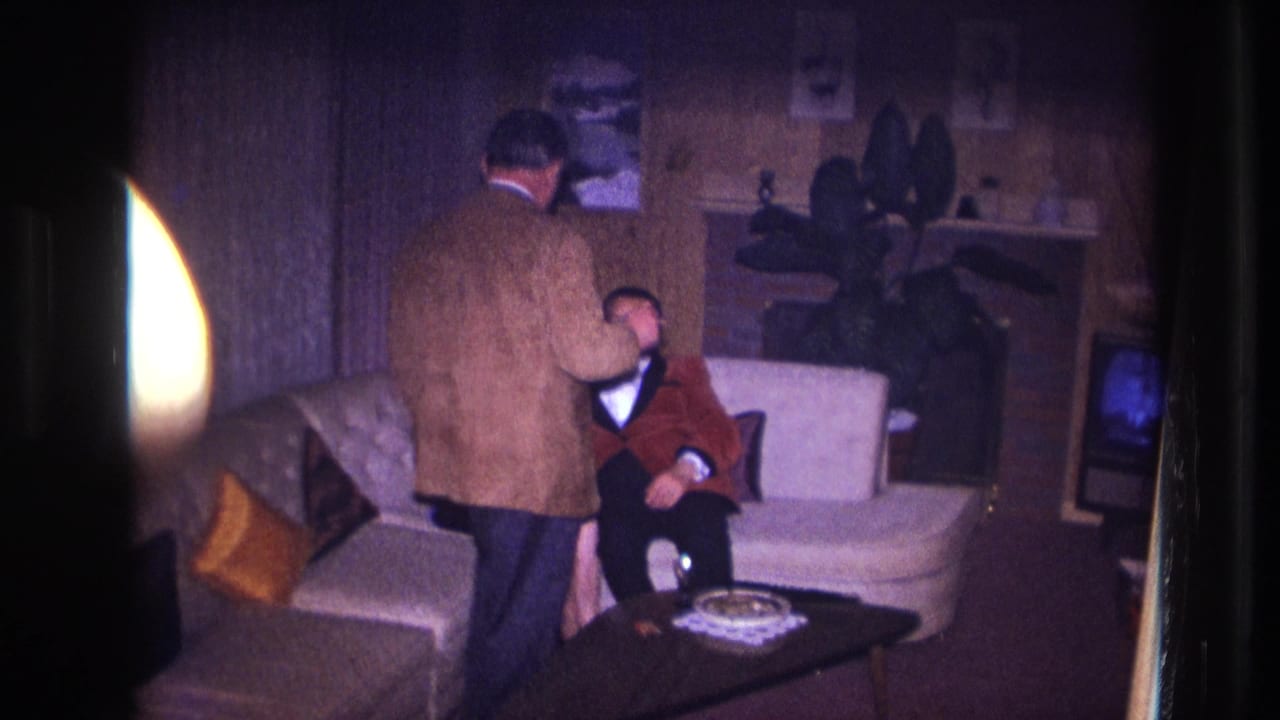 Man stands near a seated man in a red jacket beside a white couch and coffee table.