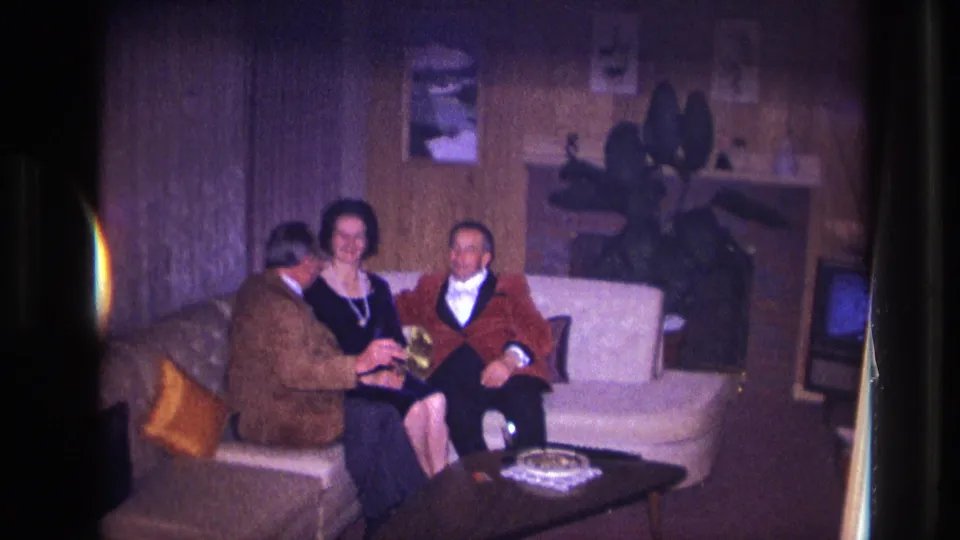 1967 Ottawa home-movie frame of three adults seated on a couch, with the man in brown holding a small object as they converse.
