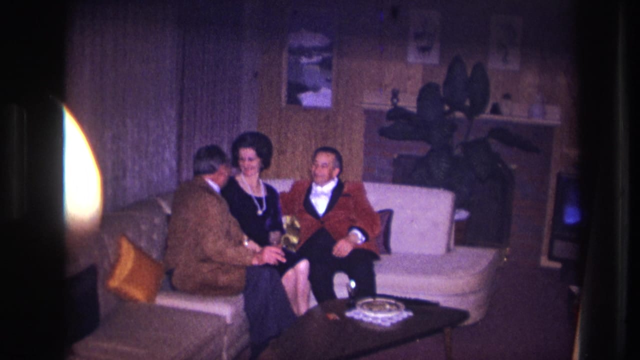 In supported 1967 Ottawa home-movie footage, two men and a woman sit together on a white sofa in a wood-paneled living room.