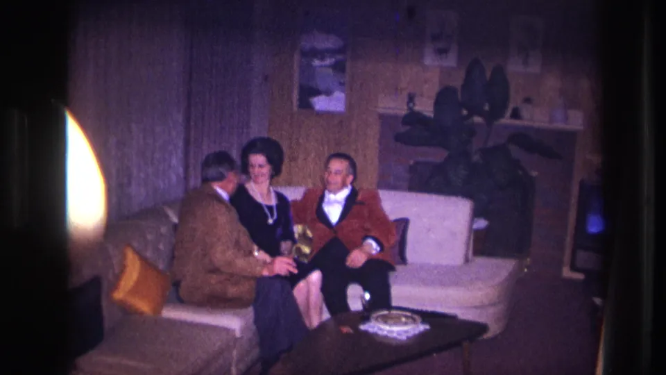 In supported 1967 Ottawa home-movie footage, two men and a woman sit together on a white sofa in a wood-paneled living room.