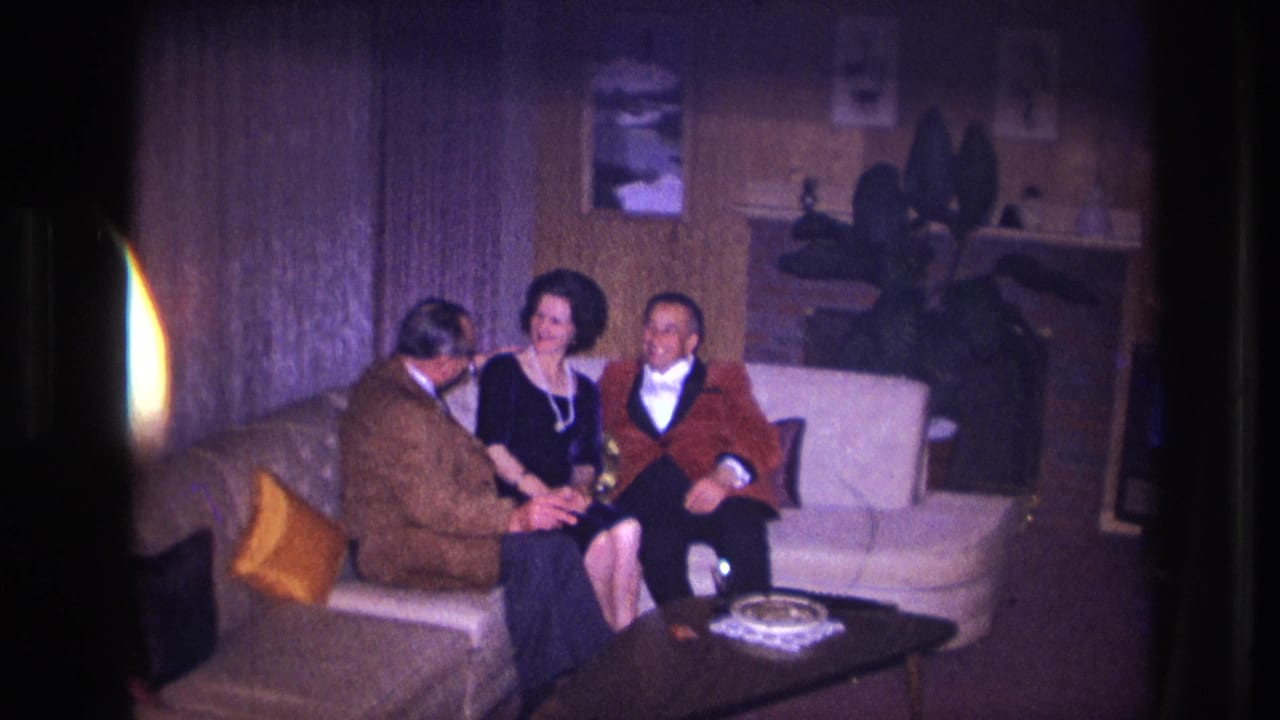 A man in a brown jacket leans toward a seated woman and man on a sofa.