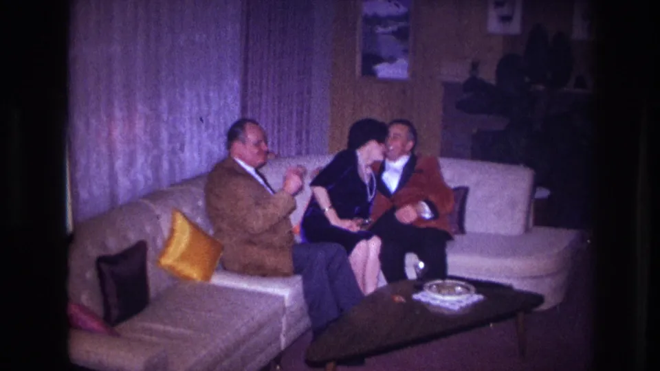 The woman in black leans forward between two seated men in the 1967 Ottawa home interior, with a coffee table in front.