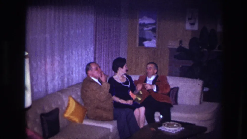 1967 Ottawa home-movie frame showing a woman seated between two men on a beige couch in a wood-paneled living room.