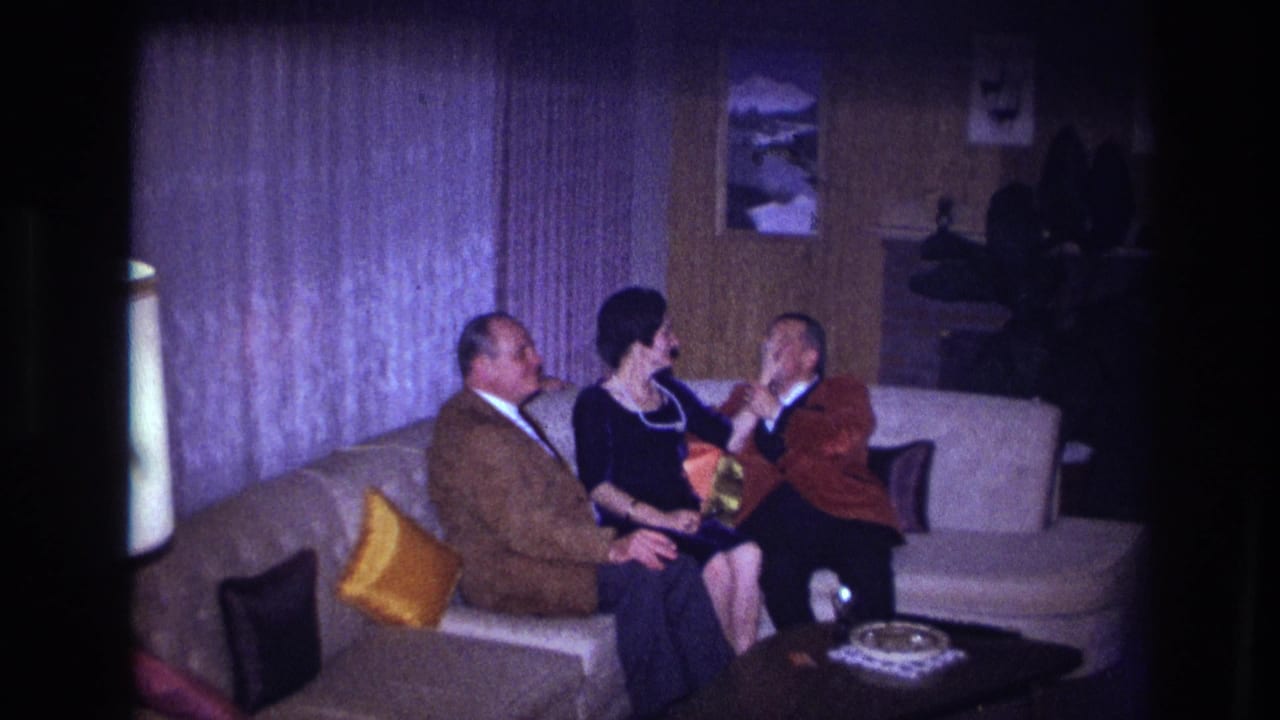1967 Ottawa home-movie frame of a woman in a black dress gesturing while seated between two men on a living-room couch.
