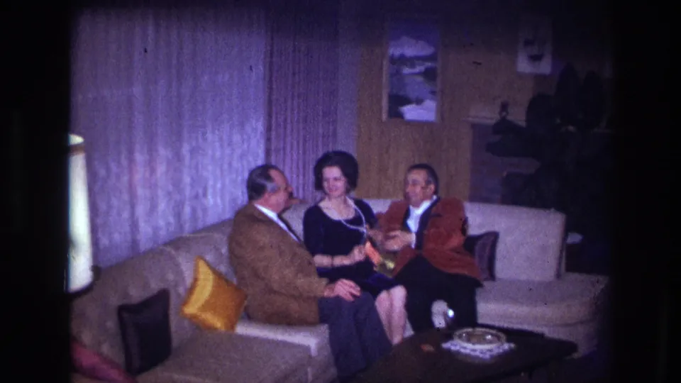 1967 Ottawa home-movie frame with three adults seated on a beige couch, the woman facing forward between the two men.