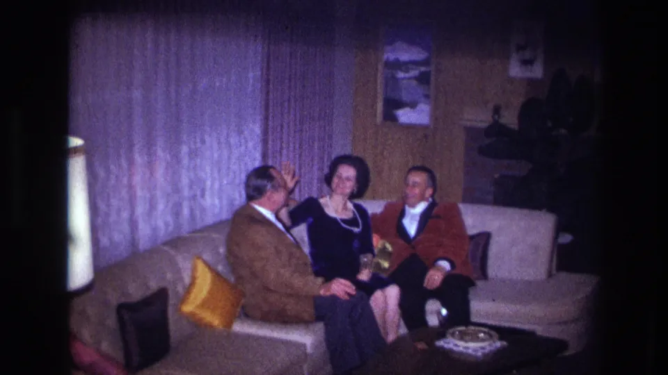 1967 Ottawa home-movie frame showing the woman raising her hand as the two men sit beside her in a private living room.