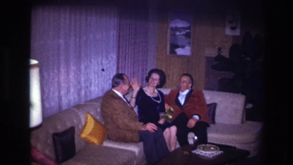 1967 Ottawa home-movie frame with a woman gesturing between two seated men in a wood-paneled living room.