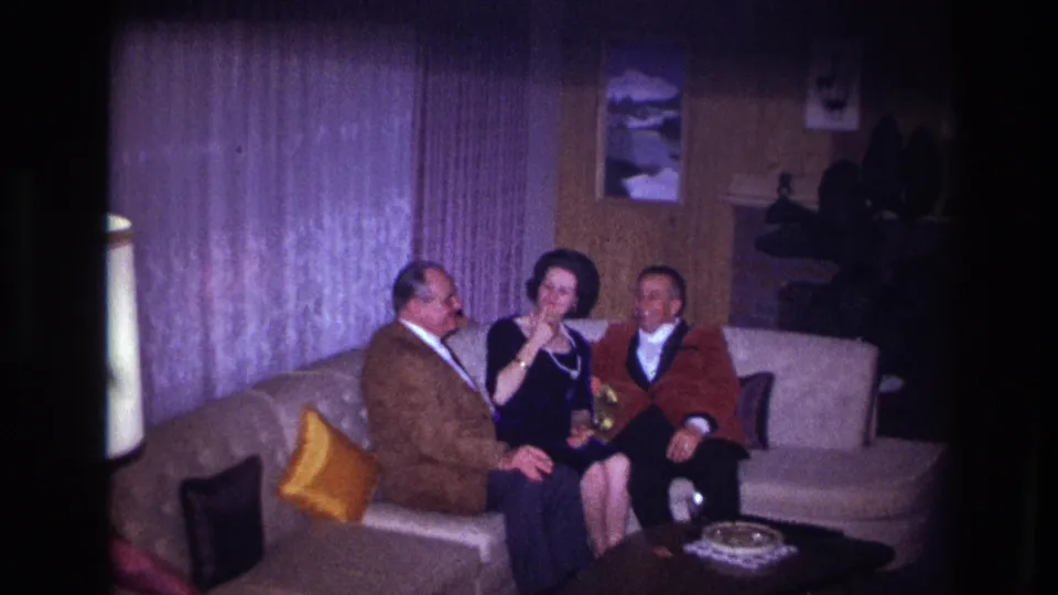 Ottawa 1967 frame with three formally dressed adults smiling on a sofa, with curtains, wall art, and lamp visible.